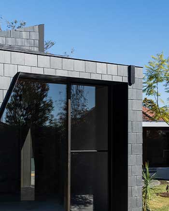 Modern building facade with dark, rectangular shingles and large glass windows reflecting trees. Minimalist design with angular lines, under a clear blue sky. Small section of a garden and part of another structure are visible in the background.