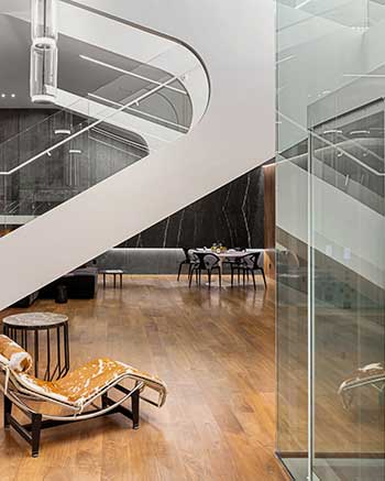 A modern interior featuring a sleek wooden floor, a stylish lounge chair with an orange patterned cushion, a round wooden side table, and a contemporary staircase with glass railings. In the background, there is a dining area with a table and chairs.