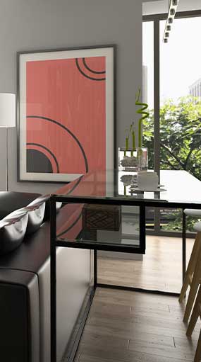 Modern living room with a glass-top desk against a large window overlooking greenery. The desk has a small sculpture and books on it. A large framed abstract artwork with red, black, and white tones hangs on the gray wall. A sleek black sofa sits nearby.