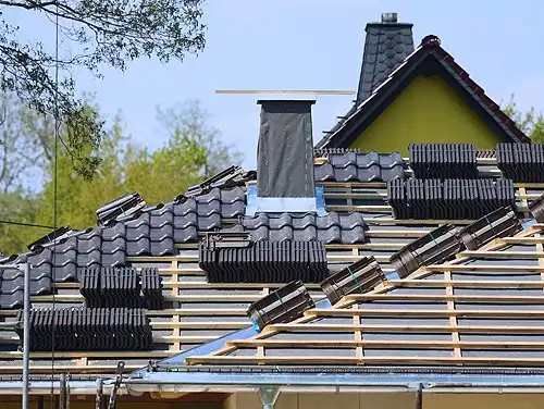 A house roof under construction with dark shingles partially installed. Stacks of shingles are placed on the wooden supportive beams. A chimney is present, and trees can be seen in the background under a clear blue sky.