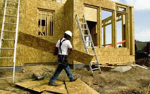 A construction worker wearing a white hard hat carries a large wooden board in front of a partially built wooden house. Two ladders are leaning against the structure, and various construction materials and tools are scattered on the ground.