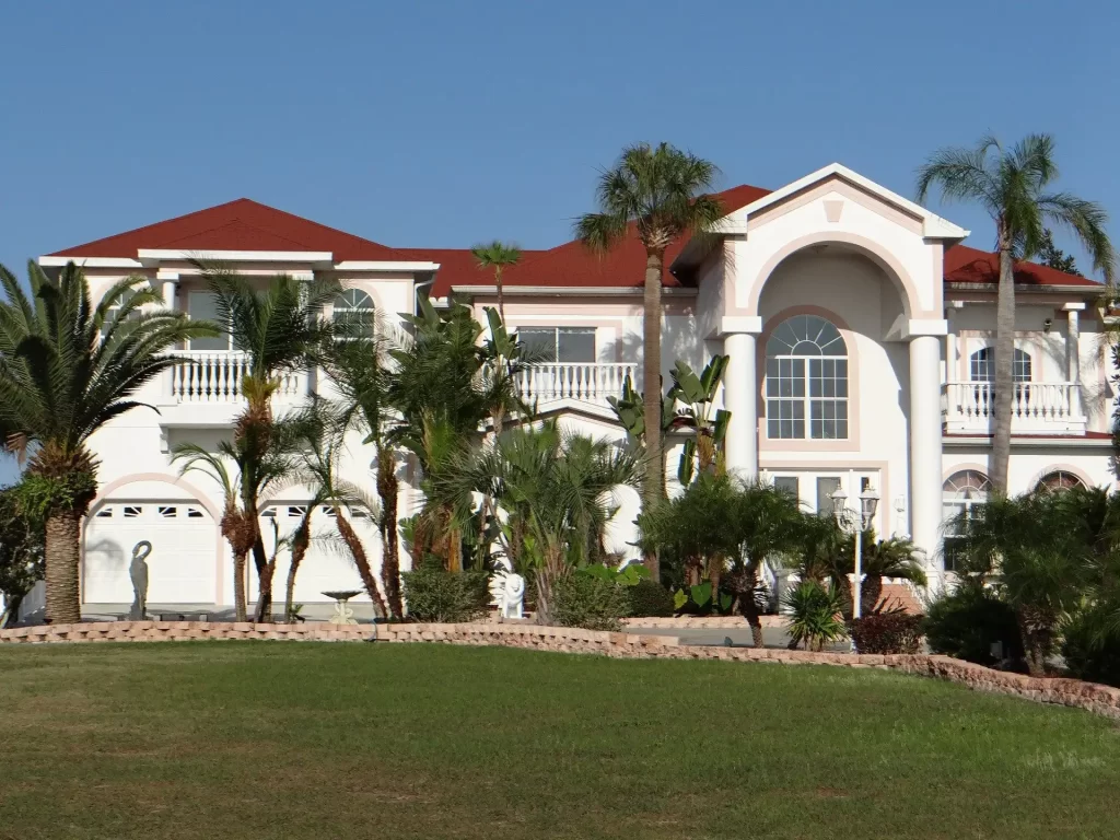 A large, luxurious two-story house with a red-tiled roof, white exterior, and multiple arched windows. The front yard features palm trees, various shrubs, and manicured grass. The sky is clear and blue in the background.