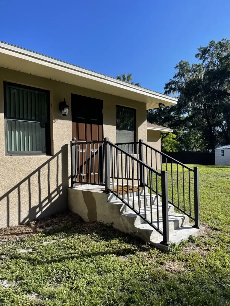 A beige house with a brown front door and black handrails on the steps leading to the entrance. The house, perfect for Mother In Law Additions in Lakeland, has a green lawn, a small window with vertical blinds next to the door, and another white building is visible in the background under a clear blue sky.