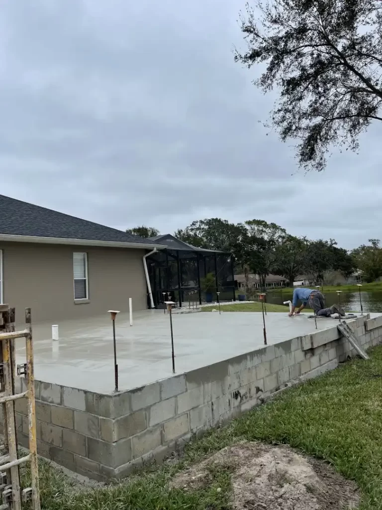 A construction worker kneels on a newly poured concrete slab adjacent to a house, smoothing the surface. The slab is elevated, with concrete blocks forming the foundation—ideal for Mother In Law Additions in Lakeland. The beige house with a dark roof features a screened-in area and overlooks a tree and lawn under a cloudy sky.