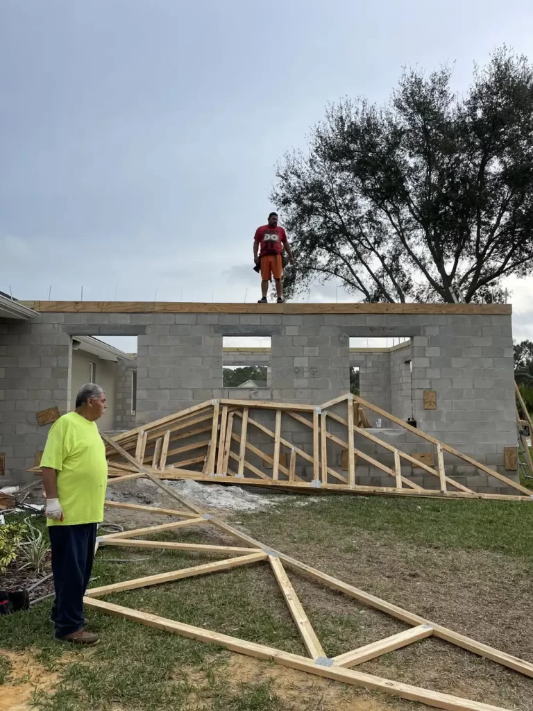 A person in a yellow shirt stands on the ground, observing another in a red shirt who is positioned on the partially-built roof of a Mother In Law Addition in Lakeland. Wooden frames and a large tree are set against a cloudy sky backdrop.