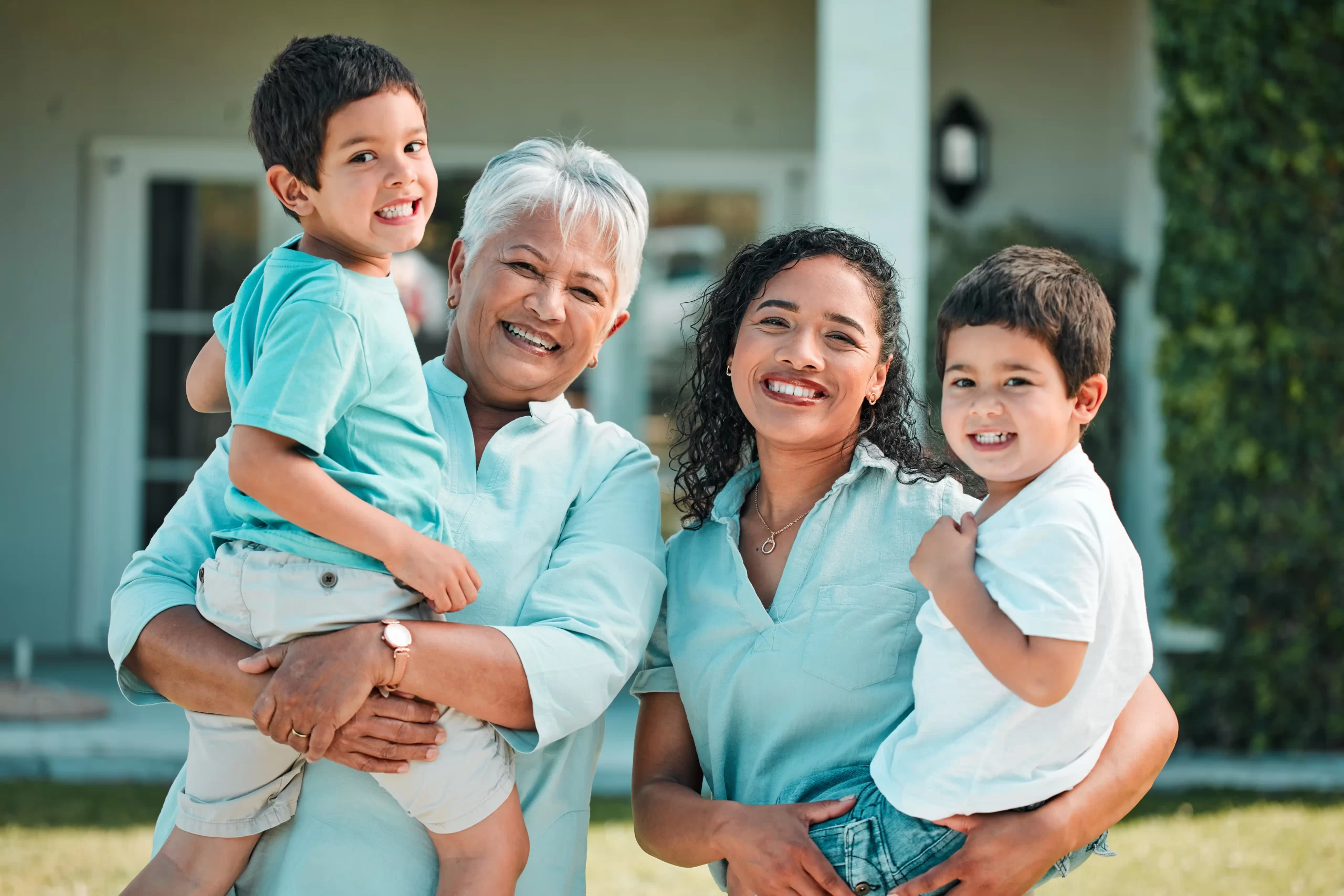 A joyful group portrait of an elderly woman, an adult woman, and two young boys stands against the charming backdrop of a Lakeland house. Both women are smiling and holding one boy each, all wearing matching light blue shirts as they bask in the warmth of their outdoor gathering.