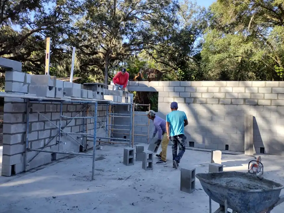 Four construction workers are building a concrete block structure outdoors in Lakeland, surrounded by trees. Two workers are on scaffoldings, laying blocks, while two others prepare materials on the ground. A wheelbarrow sits in the foreground.