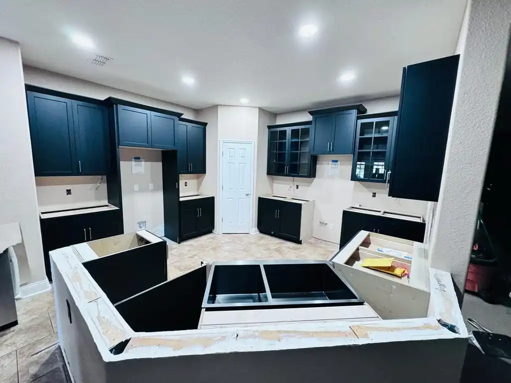 This image shows a modern Lakeland kitchen under construction. The space features dark cabinetry, with some cabinets already installed and others partially in place. Bare countertops await installation, and there is a temporary setup with various tools and materials. The floor is tiled.