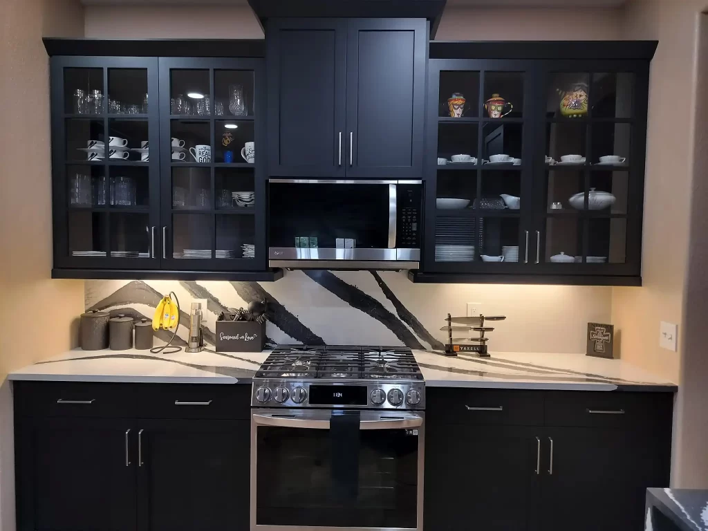 A modern Lakeland kitchen with dark cabinets featuring glass doors showcasing dishes and glassware. The backsplash has a white and black marble design. A stainless steel stove with an overhead microwave is centered. The countertop is clean, holding a coffee maker and decorative items.