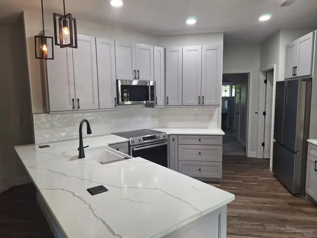 Lakeland modern kitchen with white marble countertops and backsplash, gray cabinetry, stainless steel appliances including an oven and refrigerator, a black faucet, and pendant lighting above the island. Hardwood floors run throughout the kitchen area.