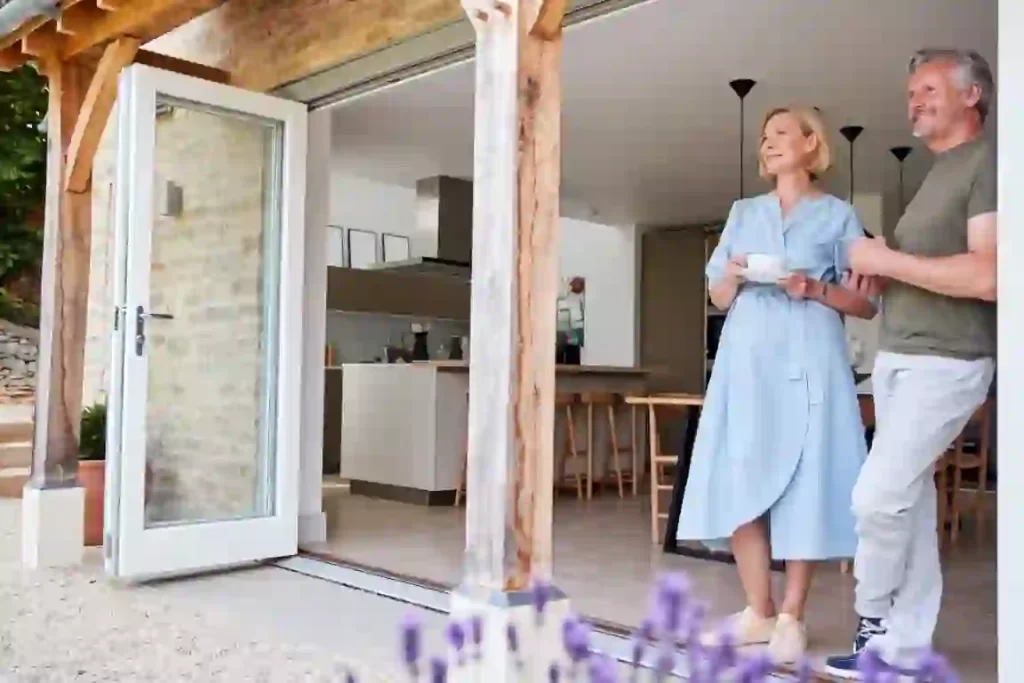 A smiling couple stands at the open doors of a modern kitchen, enjoying a view. The woman in a light blue dress holds a cup, while the man in casual clothes places a hand on her back. Wooden beams and a stone wall add rustic charm. Master the art of creating such serene spaces with key considerations in design.