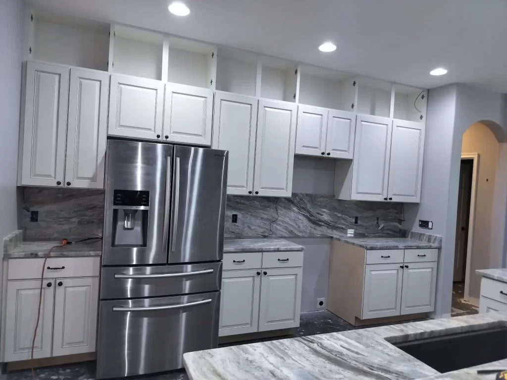 A modern kitchen under construction by Evangelisto Construction features white cabinets, a stainless steel refrigerator, marble countertops, and backsplash. Some cabinet doors are missing, and the floor appears unfinished. Recessed ceiling lights illuminate the space in this ongoing kitchen remodel.