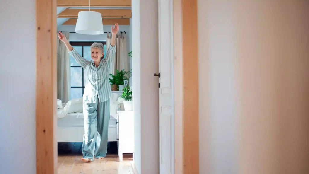 An elderly person in striped pajamas stands with arms raised in a bright bedroom with a large window and potted plants. The room, part of the new mother-in-law suite, has light-colored walls and wooden flooring, creating a serene and cozy atmosphere.