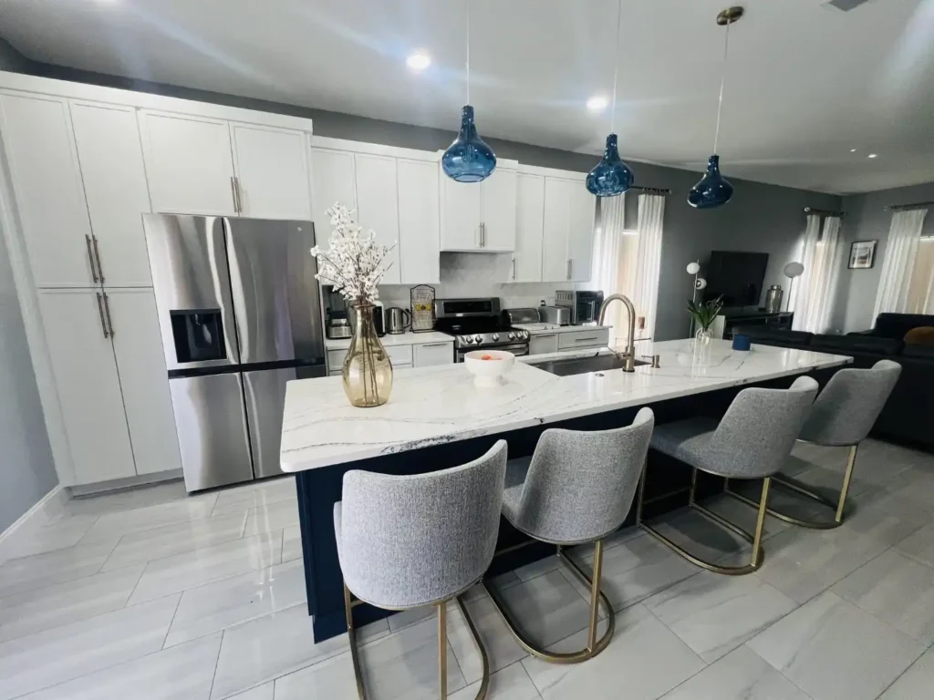 Modern kitchen renovation in Lakeland, Florida featuring white cabinets, stainless steel appliances, a marble island with sink, four gray upholstered chairs, blue pendant lights, and decorative vases. Gray tiled flooring and an open, bright layout make this space ideal for kitchen cabinet installation, home additions, or mother-in-law suite designs.