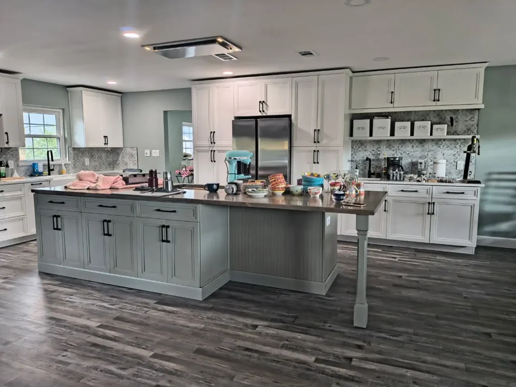 Modern kitchen renovation in Lakeland, Florida with white cabinets, stainless steel appliances, a gray island, and dark wood floors. Kitchen items and baked goods are arranged on the island countertop, with natural light streaming in from side windows—ideal for kitchen cabinet installation, home additions, or mother-in-law suite upgrades.
