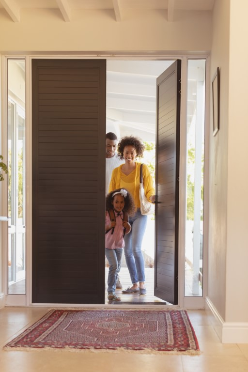 A smiling family of three, including a young girl, stands in the doorway of their home. The front door is open, sunlight streams in, and a patterned rug adds warmth—perfect for families considering Aging in Place Renovations.