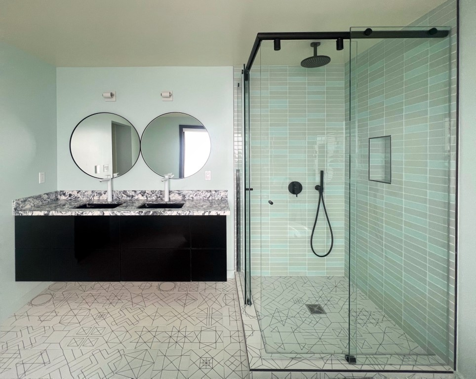 Modern bathroom with a glass-enclosed shower, light blue wall tiles, double sinks with round mirrors, black floating vanity, and geometric patterned floor tiles—ideal for stylish Aging in Place renovations.