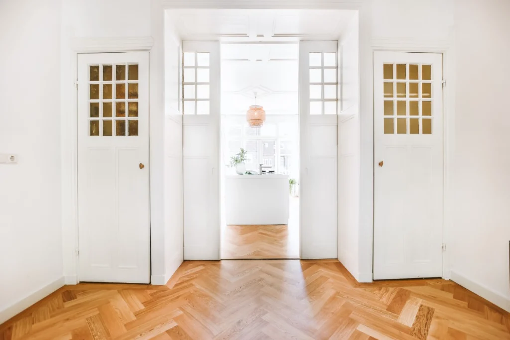 A bright, minimalist hallway with wooden herringbone flooring leads to a sunlit room. Ideal for Aging in Place Renovations, the space features wide white double doors and natural light, enhancing comfort and accessibility throughout your home.