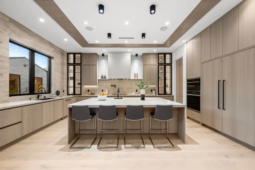 Modern kitchen renovation in Lakeland, Florida featuring light wood cabinets, a large island with four gray barstools, built-in appliances, black fixtures, and expansive windows for natural light. Minimalist decor and recessed ceiling lights make this layout ideal for kitchen cabinet installation, home additions, or mother-in-law suite conversions.