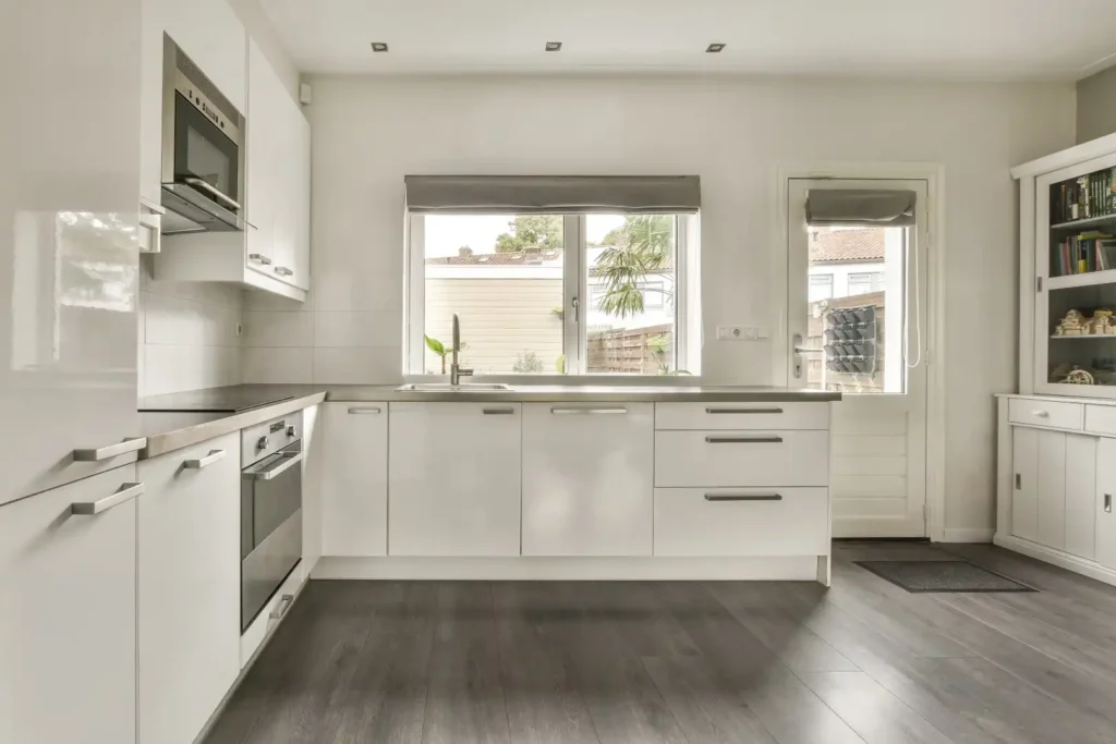 Bright modern kitchen renovation in Lakeland, Florida featuring white cabinets, stainless steel appliances, a large window, exterior door access, and light gray wood flooring. Ideal for custom kitchen cabinet installation, home additions, or mother-in-law suite designs, with a tall white cupboard providing added storage.
