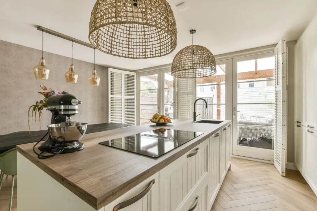 Custom kitchen cabinet installation in a modern kitchen renovation in Lakeland, Florida, featuring a wooden island with built-in cooktop, stand mixer, and fruit bowl. This spacious design suits a home addition or mother-in-law suite, with wicker pendant lights, large shuttered windows, and a dining area accented by hanging glass lamps.