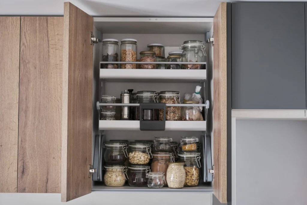 An open wooden kitchen cabinet, perfect for home renovations or aging in place upgrades, is filled with glass jars containing various dry food items such as nuts, grains, and spices, all neatly organized on three shelves.