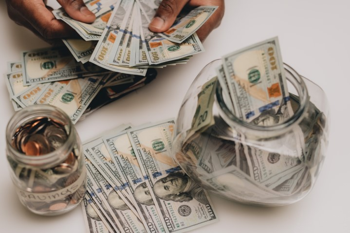 A person counts a stack of U.S. $100 bills, with more bills spread on a table—perhaps budgeting for home renovation costs. Two glass jars, one filled with coins and the other with cash, are also visible on the white surface.