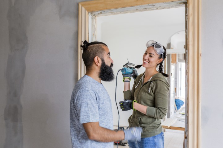 A woman wearing safety goggles and gloves holds a power drill, smiling at a bearded man as they work together on a home renovation project, carefully managing home renovation costs while standing in a doorway under construction.